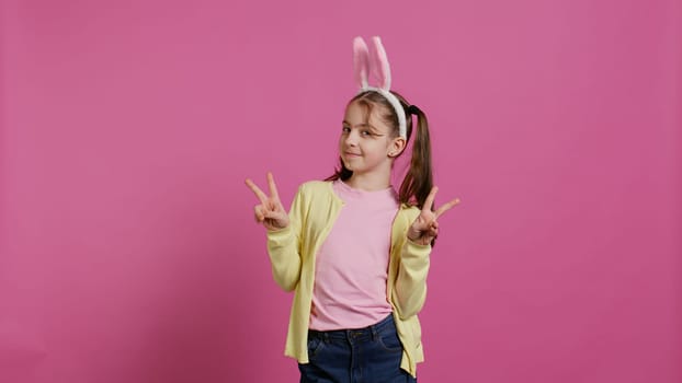 Young cheerful kid with pigtails showing peace sign in studio, feeling excited and confident about easter sunday celebration. Smiling youngster does signs against background. Camera B.