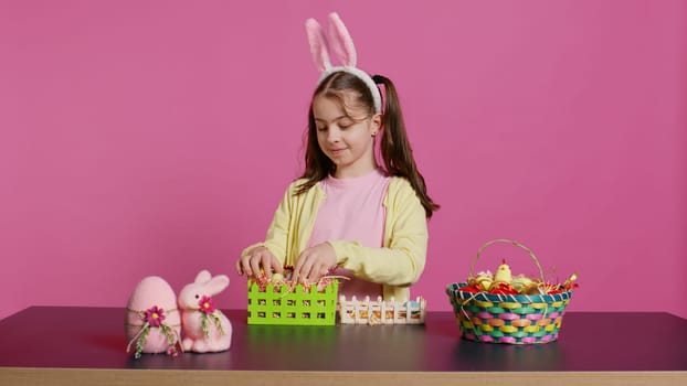 Joyful little girl showing her handcrafted festive basket on camera, creating a colorful handmade arrangement in time for easter holiday. Small toddler with bunny ears decorating eggs. Camera B.