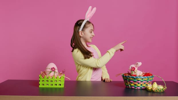 Adorable little kid indicating all directions in front of camera, pointing up, down, left and right while sitting at the table. Smiling lovely girl with bunny ears looking around. Camera B.