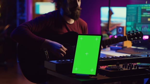 Composer performing acoustical guitar in his home studio, playing and practicing on strings. Musician learning multiple songs on instrument, uses tablet to run greenscreen display. Camera A.