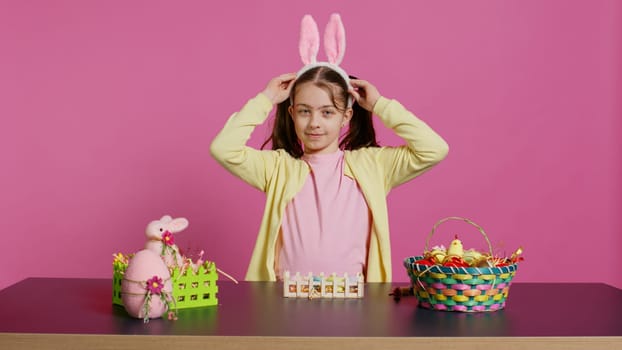 Energetic young girl with adorable bunny ears waving in studio, saying hello and greeting someone while she creates easter decorations. Joyful toddler posing against pink backdrop. Camera B.