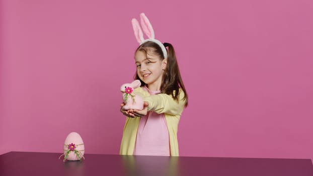 Smiling toddler with bunny ears showing her handmade easter ornaments, presenting a handcrafted decorated egg and rabbit. Young sweet schoolgirl holding festive decorations. Camera A.