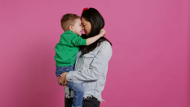 Adorable small boy hugging his mother and playing around, having fun against pink background. Young toddler posing with his mom holding him, laughing and being cheery. Camera B.