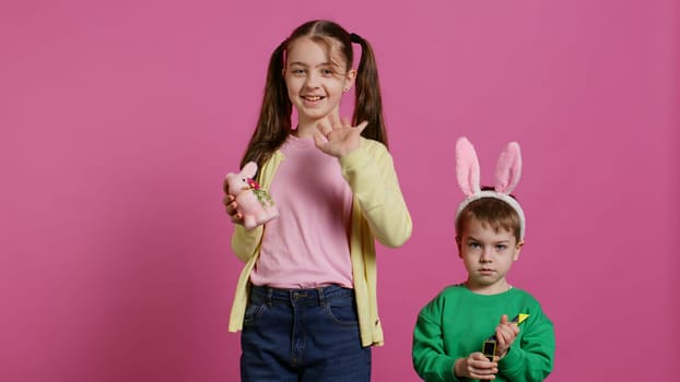 Cute brother and sister posing against pink background in studio, wearing bunny ears and playing with toys. Cheerful siblings feeling excited about easter, traditional spring holiday. Camera B.