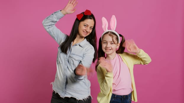 Smiling cheerful mother and little girl waving in front of camera, having fun and enjoying easter holiday celebration. Joyful schoolgirl posing with her mom in studio, saying hello. Camera B.