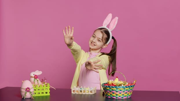 Energetic young girl with adorable bunny ears waving in studio, saying hello and greeting someone while she creates easter decorations. Joyful toddler posing against pink backdrop. Camera A.