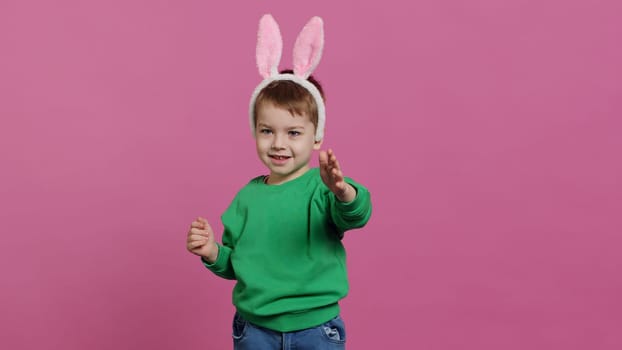 Cute little boy wearing fluffy bunny ears in studio and waving, being adorable against pink background. Smiling young kid being excited about easter holiday festivity, childhood innocence. Camera A.