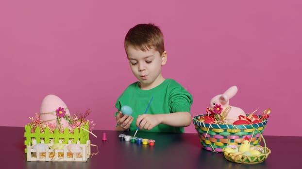 Joyful small kid painting eggs for easter holiday festivity in studio, using watercolor and art supplies. Smiling preschooler coloring festive ornaments in preparation for sunday. Camera A.