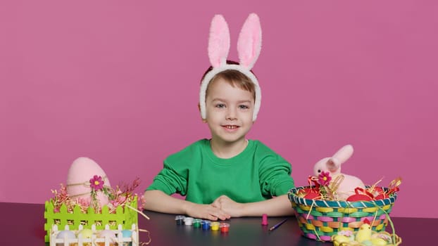 Young small kid placing bunny ears on his head in studio, preparing for easter sunday holiday celebration. Cheerful little boy sitting at a table to paint eggs and craft festive ornaments. Camera A.