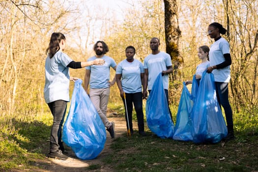 Group of nature activists collaborating to clean the woods area from trash, prepared with instruments and disposal bags. Diverse people fighting illegal dumping, ecosystem responsibility.