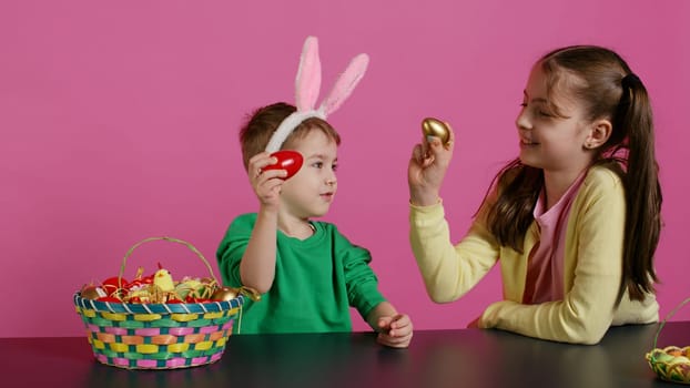 Sweet children knocking eggs together for easter tradition in studio, playing a seasonal holiday game against pink background. Lovely adorable kids having fun with festive decorations. Camera B.