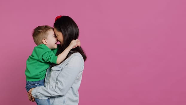 Adorable small boy hugging his mother and playing around, having fun against pink background. Young toddler posing with his mom holding him, laughing and being cheery. Camera A.