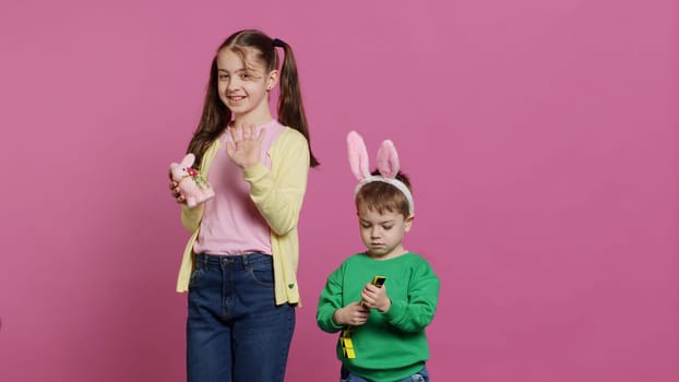 Cute brother and sister posing against pink background in studio, wearing bunny ears and playing with toys. Cheerful siblings feeling excited about easter, traditional spring holiday. Camera A.