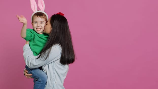 Mother lifting up her little boy and kissing him in studio, showing love and hugging cute kid against pink background. Sweet mom and son embracing each other and laughing. Camera A.