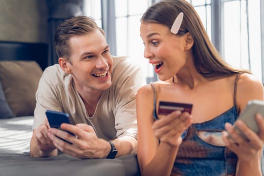 Young couple sit in the home bedroom using online payment app and digital wallet on smartphone to pay with credit card. E commerce shopping and modern purchasing via mobile internet. Unveiling