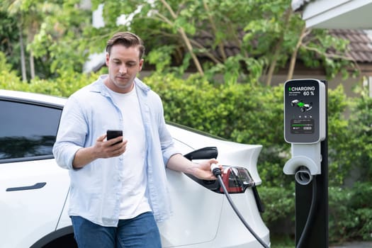 Modern eco-friendly man recharging electric vehicle from home EV charging station. Innovative EV technology utilization for tracking energy usage to optimize battery charging at home. Synchronos