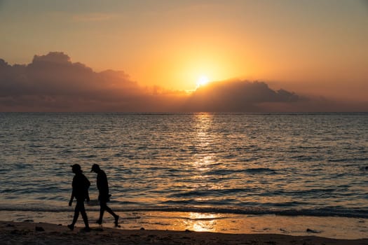 Two people walking on a beach at sunset. The sky is orange and the water is calm