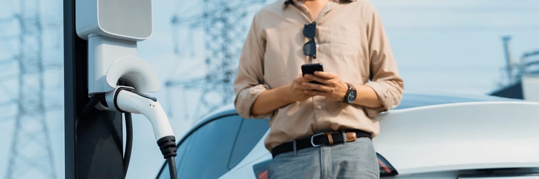 Man pay for electricity with smartphone while recharge EV car battery at charging station connected to electrical lower grid tower for eco friendly car utilization. Panorama Expedient