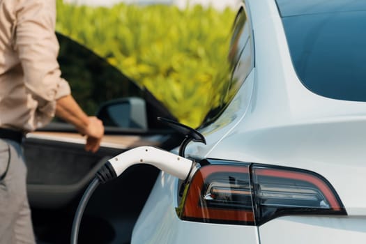 Young man recharge electric car's battery from charging station in outdoor green city park in springtime. Rechargeable EV car for sustainable environmental friendly urban travel lifestyle. Expedient