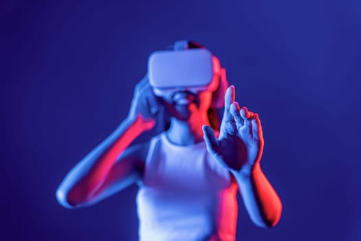 Smart female standing with surrounded by cyberpunk neon light wear VR headset connecting metaverse, futuristic cyberspace community technology. Woman using hand touching virtual object. Hallucination.