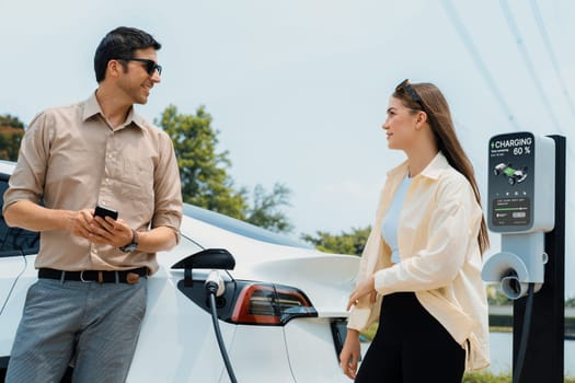 Young couple use smartphone to pay for electricity at public EV car charging station green city park. Modern environmental and sustainable urban lifestyle with EV vehicle. Expedient