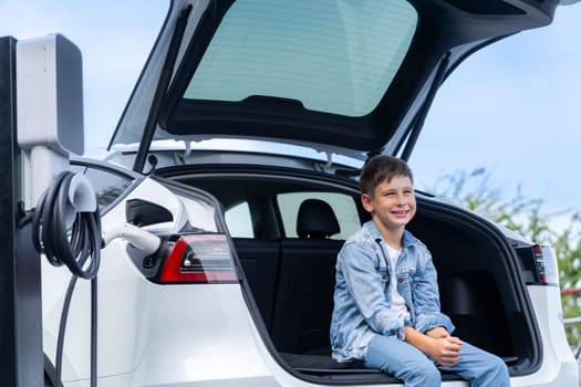 Little boy sitting on car trunk while recharging eco-friendly electric car from EV charging station. EV car road trip travel concept for alternative transportation powered sustainable energy.Perpetual