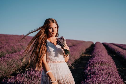 Close up portrait of young beautiful woman in a white dress and a hat is walking in the lavender field and smelling lavender bouquet.