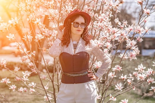 Magnolia park woman. Stylish woman in a hat stands near the magnolia bush in the park. Dressed in white corset pants and posing for the camera