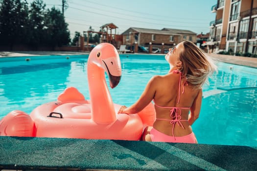 A woman in a pink bikini is sitting on a pink inflatable flamingo in a pool. The scene is playful and fun, with the woman enjoying her time in the water