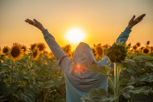 Woman is standing in a field of sunflowers, with the sun in the background. The person is holding their arms up in the air, as if they are celebrating or expressing joy