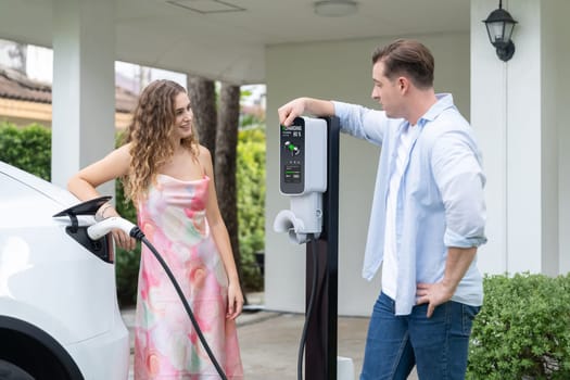 Happy and lovely couple with eco-friendly conscious recharging electric vehicle from EV home charging station. EV car technology utilized for residential home to future sustainability. Synchronos