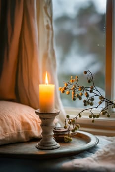 A candle is lit in a window, with a branch of berries next to it. The candle is on a small white pedestal, and the berries are on a tray. The scene is cozy and inviting. Generative AI