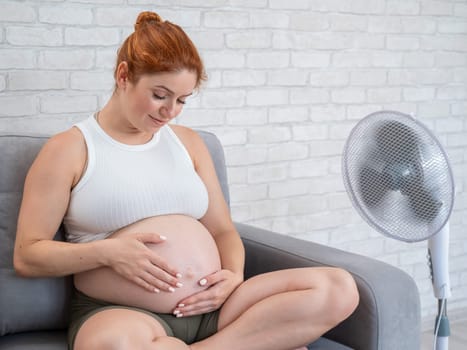 A pregnant woman is sitting on the couch, stroking her tummy and enjoying the cool air from an electric fan