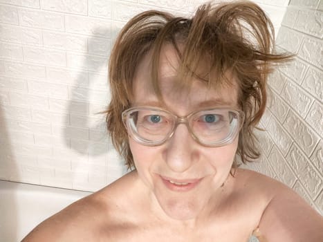 Shaggy cheerful middle aged woman with glasses smiling in bathroom in early morning. Smiling Woman Relaxing and taking selfie in mirror