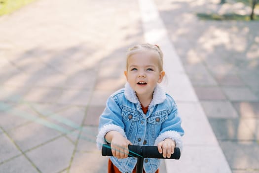 Little smiling girl rides a scooter leaning on the steering wheel and looking up. High quality photo