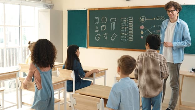 Diverse elementary student walking in classroom while teacher looking at child. Cute children attend classroom while professional instructor prepare for teaching and standing at blackboard. Pedagogy.