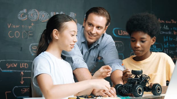 Caucasian teacher looking at laptop screen while explain car model system at blackboard with coding or programing code written. Happy diverse student learning about robotic construction. Edification.