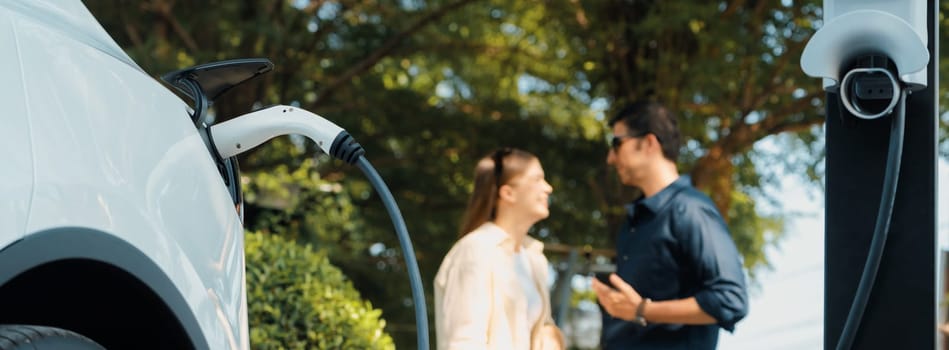 Young couple recharge electric car battery from charging station in green city park in springtime. Rechargeable EV car for sustainable environmental friendly urban travel lifestyle. Panorama Expedient