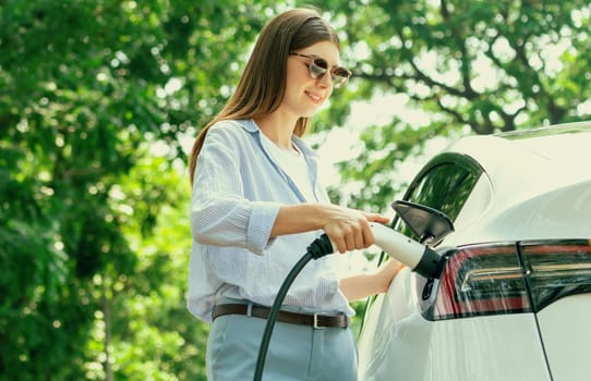 Young woman recharging battery for electric car during road trip travel EV car in natural forest or national park. Eco friendly travel during vacation and holiday. Exalt