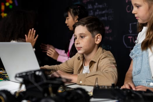 In classroom student in STEM class learning about coding robotics car. Brown shirt Schoolboy and white bib schoolgirl watching laptop and discussing. Other schoolgirls playing around funny. Erudition.