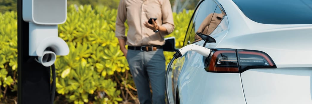 Young man use smartphone to pay for electricity at public EV car charging station green city park. Modern environmental and sustainable urban lifestyle with EV vehicle. Panorama Expedient