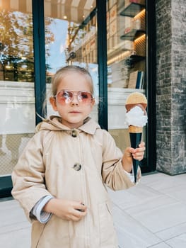 Little girl in sunglasses posing with a big ice cream cone in her hand near the glass door of the building. High quality photo