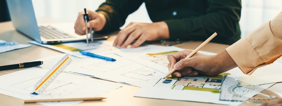 A cropped image of professional engineer using divider to measure blueprint at meeting table with blueprint, laptop and architectural equipment scatter around. Closeup. Delineation.