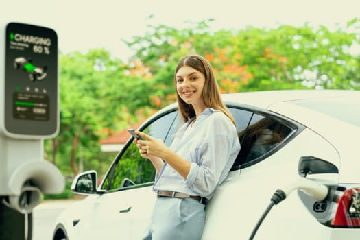 Young woman using smartphone online banking application to pay for electric car battery charging from EV charging station during vacation holiday road trip at national park or summer forest. Exalt