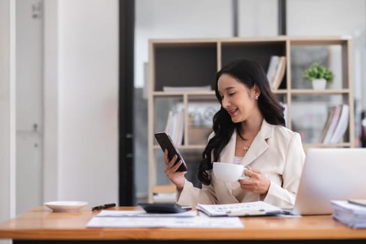 Beautiful business woman explores social media on her phone and drinks coffee before starting work in the morning at her desk..