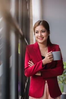 Confident smiling business woman stands holding a coffee mug and looks out the window..