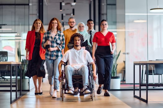 A diverse group of young business people walking a corridor in the glass-enclosed office of a modern startup, including a person in a wheelchair and a woman wearing a hijab.