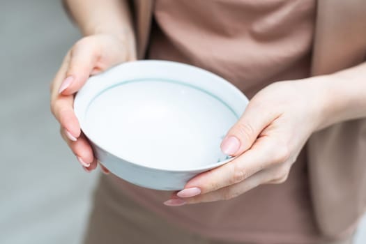 Unrecognizable woman holding empty bowl in her hands, kitchen interior, copy space, closeup shot, cropped of hungry lady starving. Famine, poverty, hunger, crisis concept. High quality photo