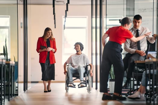 A business leader with her colleague, an African-American businessman who is a disabled person, pass by their colleagues who work in modern offices.