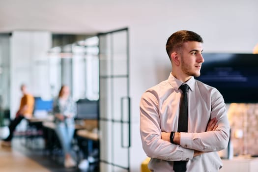 A young business leader stands with crossed arms in a modern office hallway, radiating confidence and a sense of purpose, embodying a dynamic and inspirational presence.